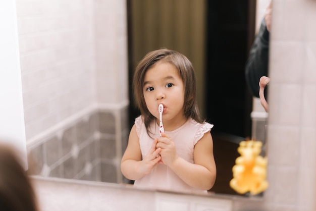 Adorable y hermosa niña se está cepillando los dientes con un cepillo de dientes frente al espejo del baño, mirando a la cámara.