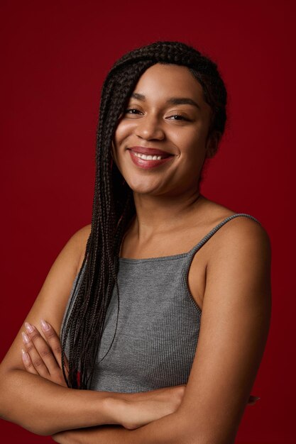 Adorable joven afroamericana con largas trenzas rastas en camiseta deportiva gris sonriendo linda sonrisa con dientes mientras mira a la cámara, posando sobre fondo de color rojo con espacio publicitario de copia