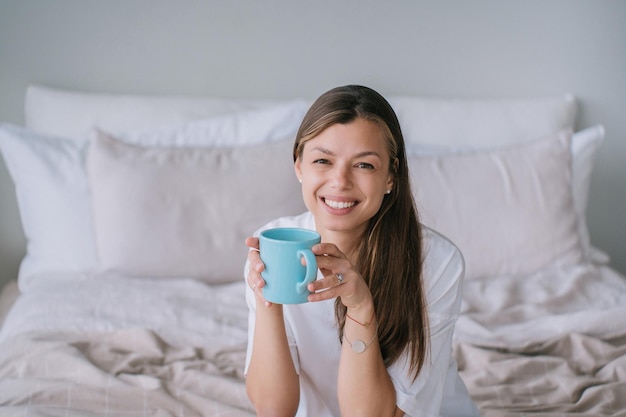 Adorable kaukasische junge Frau sitzt auf dem Bett und hält eine Tasse Tee mit Blick auf die Kamera