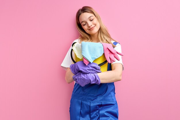 Adorable mujer caucásica en uniforme de trabajo azul de pie con cubo y trapos posando aislado en la pared rosa