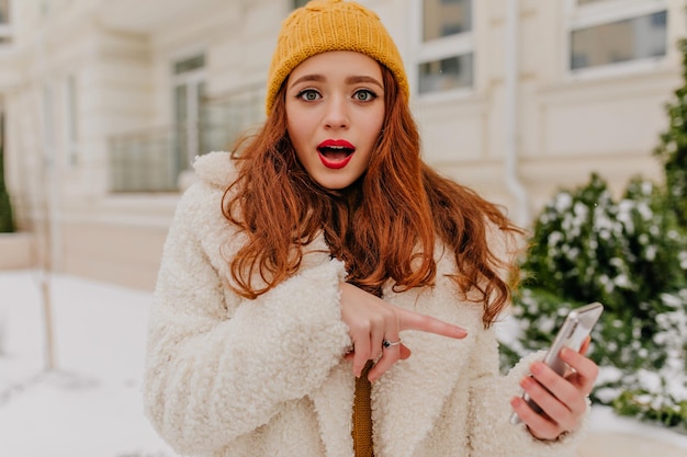 Adorable mujer europea posando con el teléfono en el día de invierno Foto al aire libre de una elegante chica pelirroja sosteniendo un teléfono inteligente
