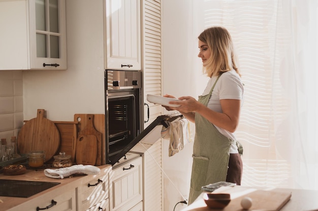 Adorable mujer joven en delantal de chef pone pastel de manzana en el horno