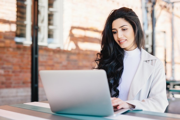 Adorable mujer morena vestida de blanco sentada al aire libre en el café usando su computadora portátil comunicándose con amigos en línea usando conexión gratuita a Internet Concepto de tecnología de estilo de vida de personas