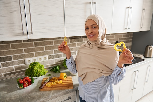 Adorable mujer musulmana en hijab lindo sostiene trozos de pimiento amarillo en su mano y sonríe a la cámara mientras cocina ensalada vegana