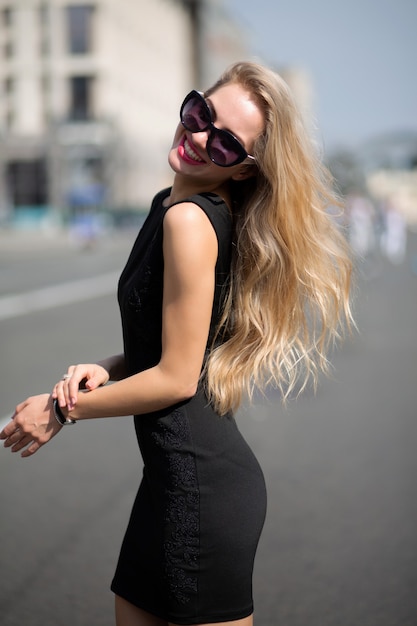 Adorable mujer rubia sonriente con vestido negro y gafas de sol, caminando por la calle soleada