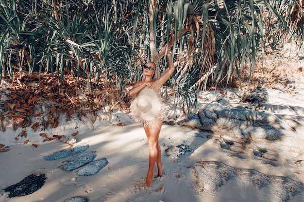 Adorable mujer sexual desnuda sin ropa escondida detrás de un sombrero de paja y posando de cuerpo entero en la arena de la playa con el telón de fondo de palmeras y vegetación. Concepto de cuerpo perfecto
