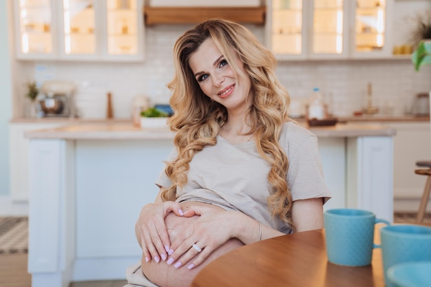 Adorable mujer sueca rubia embarazada sentada en la mesa en la cocina formando el corazón con las manos en el vientre