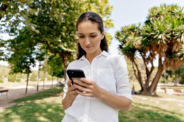 Adorable mujer del vestíbulo con el pelo largo en camisa blanca caminando en un parque verde de verano con un smartphone.