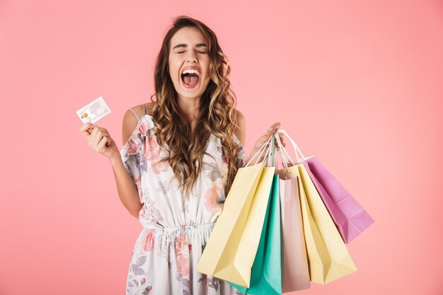 Adorable mujer vestida con tarjeta de crédito y coloridas bolsas de la compra, aislado en rosa