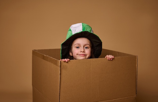 Adorable niña de 4 años con un sombrero Leprechaun se asoma por debajo de una caja de cartón y sonríe lindamente a la cámara Happy St Patrick's Day Studio filmada sobre fondo beige con espacio para copiar