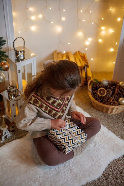 Foto adorable niña de 5 años con cabello largo y rizado y ojos azules abre cajas de regalos cerca del árbol de navidad fiestas año nuevo las vacaciones de invierno
