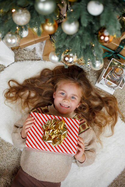 Foto adorable niña de 5 años con cabello largo y rizado y ojos azules abre cajas de regalos cerca del árbol de navidad fiestas año nuevo las vacaciones de invierno