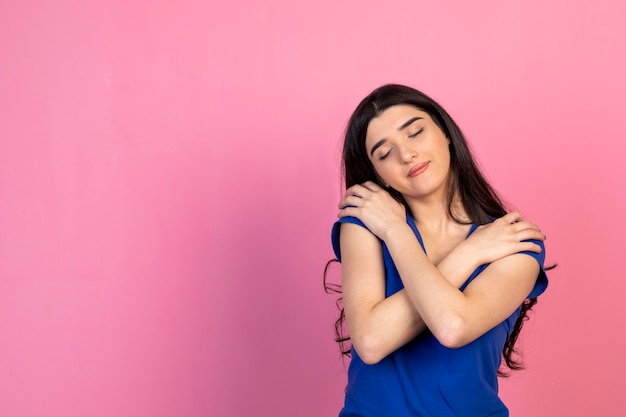 Foto adorable niña abrazándose a sí misma y de pie sobre fondo rosa