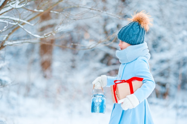 Adorable niña con abrigo cálido al aire libre el día de Navidad con regalo y linterna