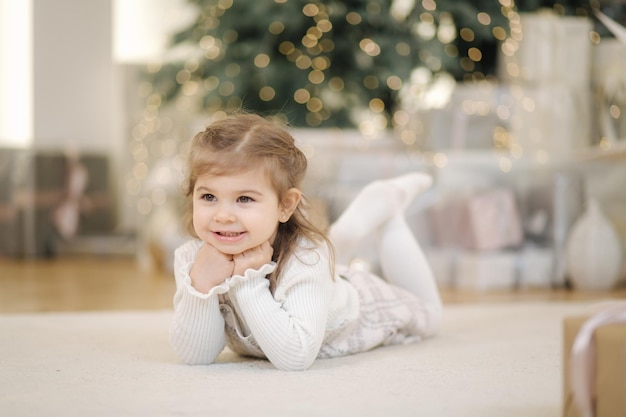 Adorable niña acostada de frente cerca de abeto y sonrisa de humor navideño en casa con una gran cantidad de