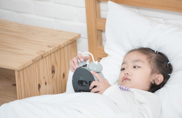 Adorable niña asiática acostado en la cama blanca mano que sostiene el despertador