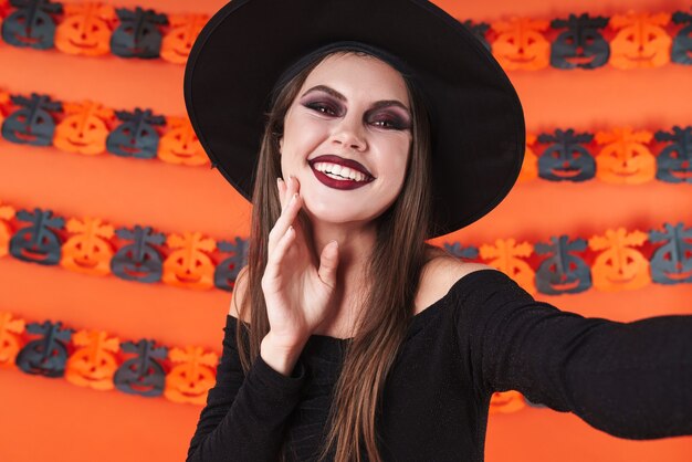 Adorable niña bruja en traje de halloween negro sonriendo a la cámara aislada sobre la pared de calabaza naranja