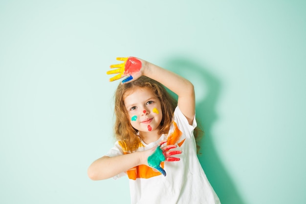 Adorable niña con cabello castaño rizado y puntos coloridos en su rostro mostrando sus palmas pintadas sobre fondo azul claro