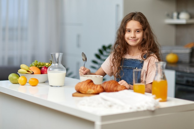 Adorable niña con cabello rizado comiendo cereales desayuno saludable en la cocina, mirando a la cámara con una cuchara en la mano.