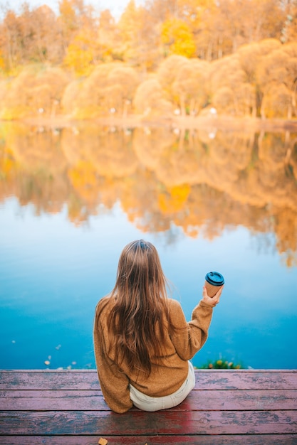 Adorable niña con café para llevar caliente al aire libre en el día de otoño cerca del lago