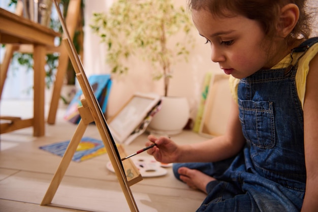 Adorable niña caucásica linda niña sentada en el suelo en un caballete de madera y dibujando pintura sobre lienzo Clase de arte creatividad concepto de entretenimiento infantil