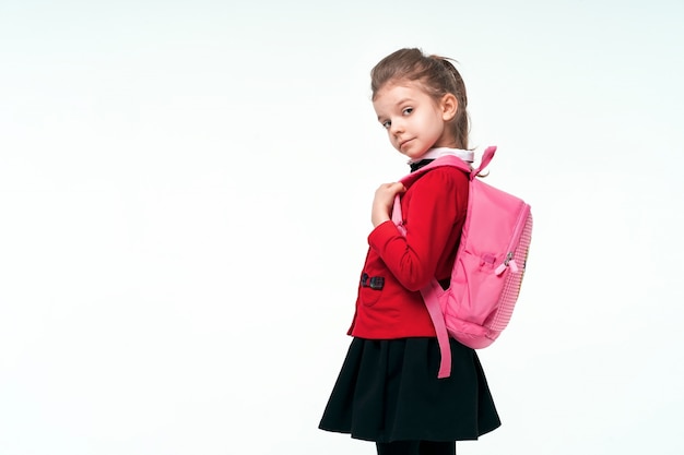 Foto adorable niña con chaqueta escolar roja, vestido negro, en las correas de una mochila y sonriendo