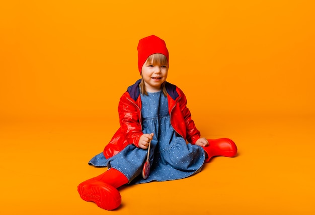 Foto adorable niña en una chaqueta roja y botas sosteniendo una paleta sobre un fondo amarillo. concepto de otoño.