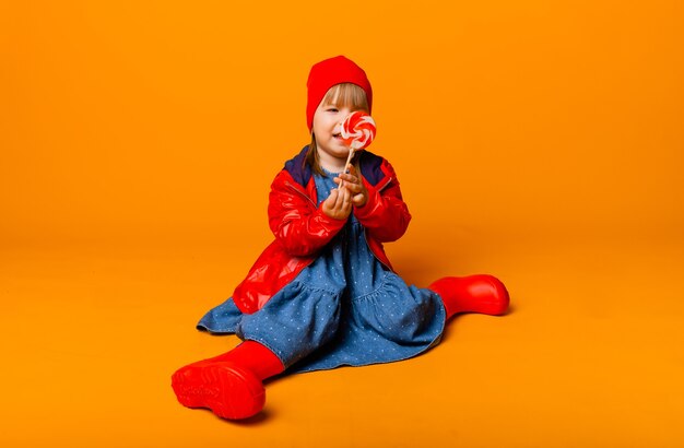 Adorable niña en una chaqueta roja y botas sosteniendo una paleta sobre un fondo amarillo. concepto de otoño.