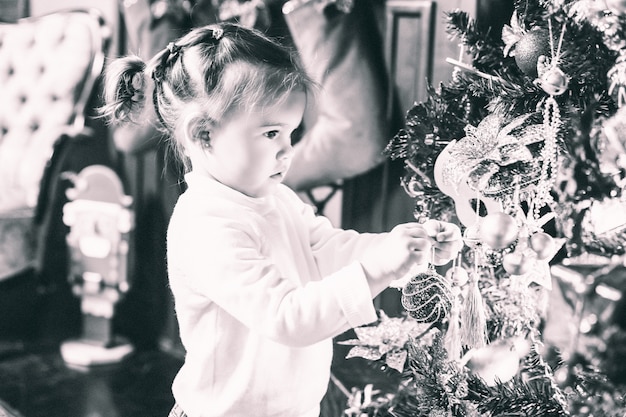 Adorable niña con colas en suéter blanco mirando el árbol de Navidad bellamente decorado