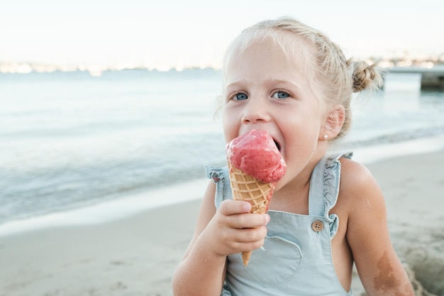 Adorable niña comiendo helado dulce y mirando a la cámara mientras se sienta en la playa de arena cerca del mar ondulante