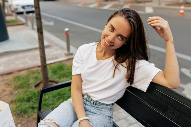 Adorable niña encantadora con ojos azules y cabello oscuro suelto con camiseta blanca y jeans descansando afuera en un banco y jugando con ella