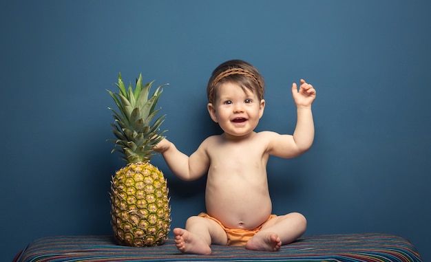 Adorable niña feliz sentada jugando con una piña sobre un fondo azul.