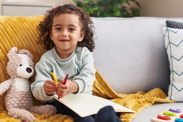 Adorable niña hispana dibujando en un cuaderno sentado en un sofá en casa