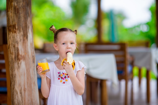 Adorable niña con jugo y plátano en el café al aire libre