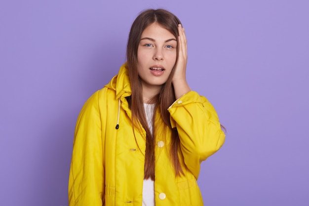 Adorable niña con la mano en la cabeza, olvidó algo importante, vestida con chaqueta amarilla, mira a la cámara con la boca abierta, tiene un error, posando aislada sobre fondo lila.