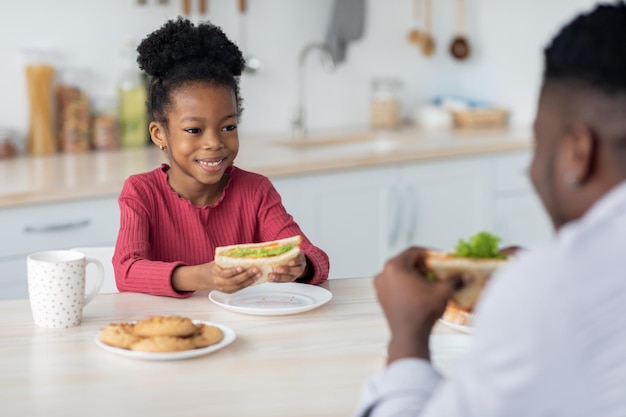 Adorable niña negra mirando a su padre mientras come un sándwich