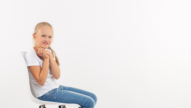 Adorable niña niño con largo cabello rubio sentado en la silla sobre fondo blanco.