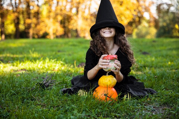 Adorable niña con el pelo rizado en ropa de bruja negra y sombrero haciendo muecas, sosteniendo algunas calabazas, sentada sobre un césped, sobre un fondo de día soleado de otoño.