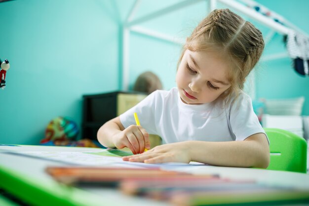 Adorable niña pequeña dibujando con lápices en casa sentado en la mesa. Niño creativo sentado en una habitación aprendiendo a dibujar. Niña pequeña haciendo los deberes en casa.