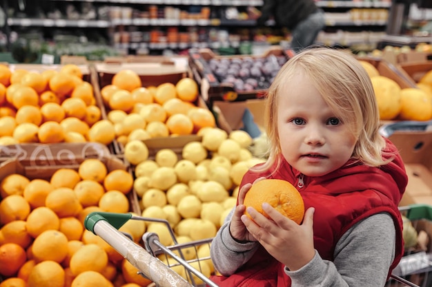 Adorable niña pequeña sentada en un carrito de compras en una tienda de frutas o en un supermercado