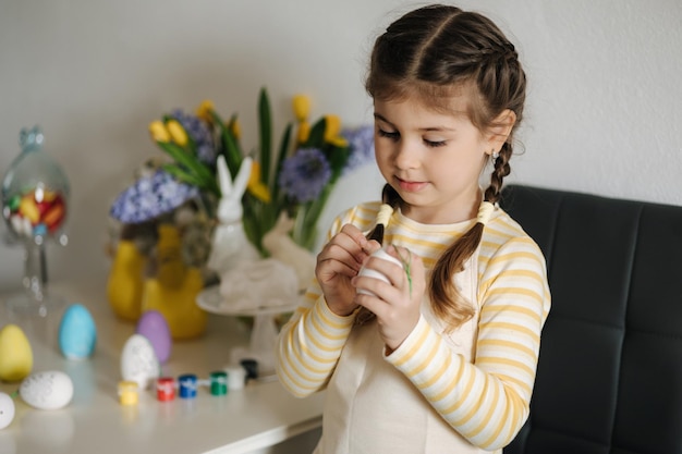 Adorable niña pintando huevos en la cocina en casa Linda chica en delantal Fondo de decoración de Pascua