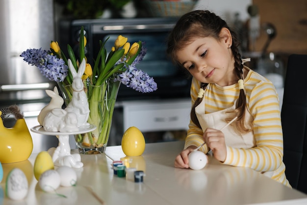 Adorable niña pintando huevos en la cocina en casa niña linda en delantal fondo de Pascua