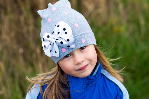 Adorable niña posando en superficie borrosa y sonriendo a una cámara. Vistiendo abrigo de invierno y sombrero. Hermosa joven en el otoño al aire libre.