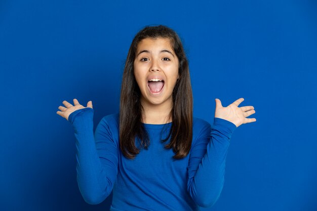 Adorable niña preadolescente con jersey azul gesticulando sobre la pared azul