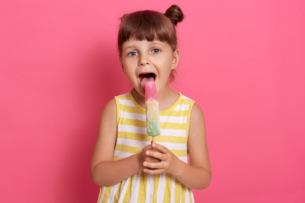 Adorable niña preescolar sosteniendo sorbete y mirando a cámara. Niña linda lamiendo helados de frutas, abre ampliamente la boca, de pie contra la pared rosa,