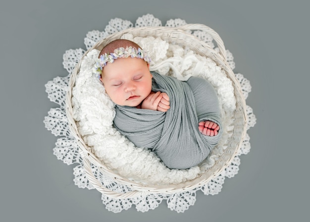 Adorable niña recién nacida con guirnalda acostada de espaldas en la canasta y durmiendo. Dulce envuelto en tela para bebés y niños que duermen la siesta durante la sesión de fotos de estudio con decoración