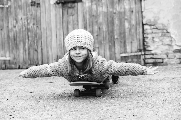 Adorable niña en ropa de lana de invierno acostado patineta vuela como un avión al aire libre en el patio trasero Sonriente niña jugando con patineta Versión vintage en blanco y negro