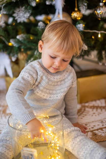 Adorable niña sentada cerca del árbol de navidad con luces festivas y regalos de navidad navidad y año nuevo