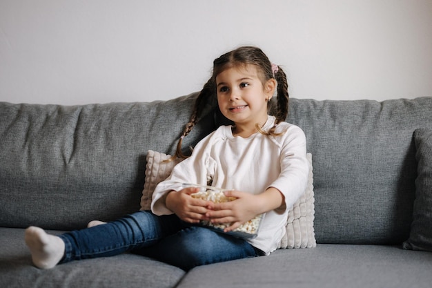 Adorable niña sentada en el sofá y viendo la televisión en casa Linda niña comiendo palomitas de maíz Estado de ánimo de vacaciones