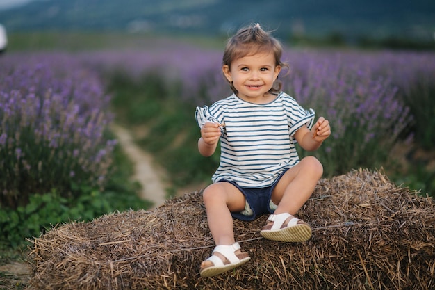 Adorable niña se sienta en el heno junto a la granja Fondo del campo de lavanda de verano Linda chica en camiseta a rayas y pantalones cortos azules